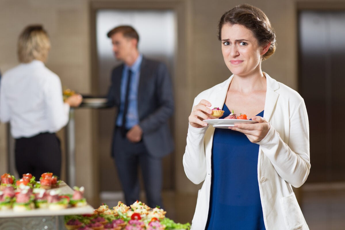 Domino's Closeup of looking at camera disgusted beautiful middle-aged woman tasting snacks from plate and standing at table with food at buffet reception with blurry people in background